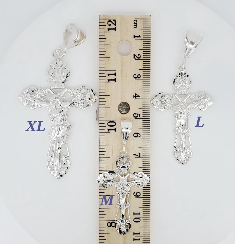 May include: Three silver filigree cross pendants of different sizes, labelled "XL", "M", and "L", are shown next to a ruler. The ruler is marked in inches and centimetres.