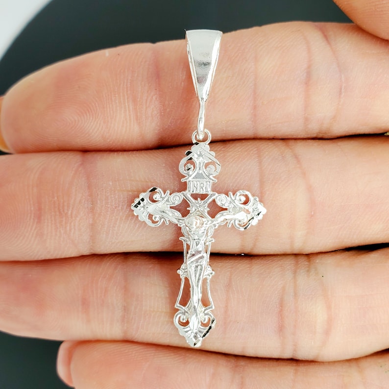 May include: A silver filigree cross pendant with a detailed design and a figure of Jesus Christ on the front. The pendant has a bail for attaching to a chain.