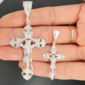 May include: Two silver filigree cross pendants with a crucifix design. The larger cross is approximately 5 cm tall and the smaller cross is approximately 2.5 cm tall.