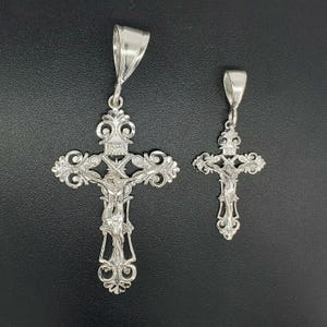 May include: Two silver crucifix pendants with intricate designs. The larger pendant features a detailed figure of Jesus Christ on the cross. Both pendants have a bail for attaching to a chain.