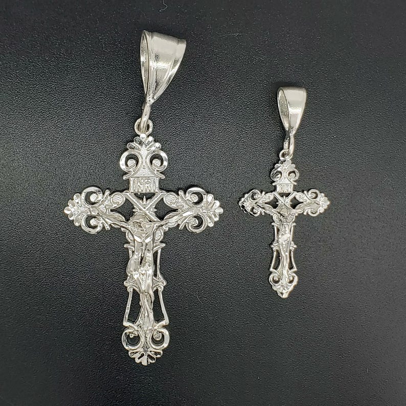 May include: Two silver crucifix pendants with intricate designs. The larger pendant features a detailed figure of Jesus Christ on the cross. Both pendants have a bail for attaching to a chain.