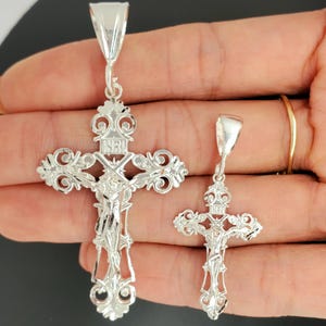 May include: Two silver crucifix pendants with intricate designs. The larger pendant has a more detailed design with a figure of Jesus Christ on the cross. The smaller pendant has a simpler design with a figure of Jesus Christ on the cross.
