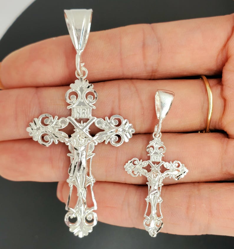 May include: Two silver crucifix pendants with intricate designs. The larger pendant has a more detailed design with a figure of Jesus Christ on the cross. The smaller pendant has a simpler design with a figure of Jesus Christ on the cross.