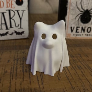 May include: White ghost-shaped Halloween decoration with cut-out eyes. The ghost is sitting on a wooden surface.