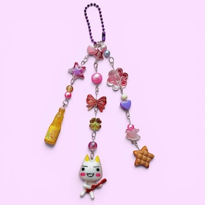 May include: A decorative charm featuring a variety of colorful beads, stars, flowers, and a small bottle. The charm also includes a cartoon cat playing a guitar and a purple chain for hanging. The charm is set against a light pink background.