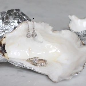 May include: A white oyster shell jewellery dish with silver trim, holding a pair of teardrop earrings and two diamond rings. The dish is set on a white surface, with two additional silver-covered oyster shells nearby.