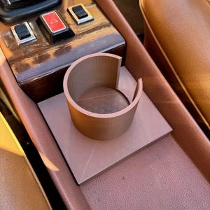 Può includere: Un interno di un'auto in pelle marrone con un portabevande marrone e una base quadrata. Il portabevande è in legno e ha una parte superiore arrotondata.