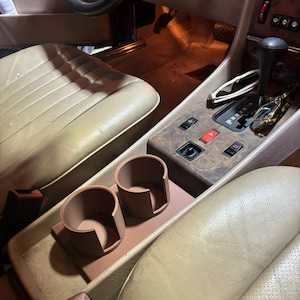 Op de afbeelding: Een bruin lederen auto-interieur met een op maat gemaakte bekerhouder voor twee drankjes. De bekerhouder is gemaakt van bruin plastic en bevindt zich in de middenconsole.