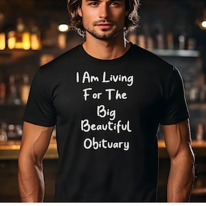 Könnte beinhalten: Schwarzes T-Shirt mit dem weißen Text "I Am Living For The Big Beautiful Obituary". Das kurzärmelige Shirt scheint aus einem weichen Material zu sein. Der Hintergrund ist verschwommen, was auf eine Bar oder ein Restaurant hindeutet.