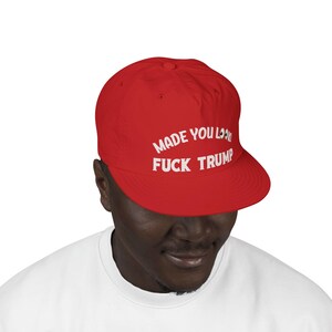 May include: A red baseball cap with the text "MADE YOU LOOK FUCK TRUMP" in white. The cap is worn by a person wearing a white shirt.