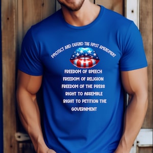 Puede incluir: Una camiseta azul real de manga corta con un gráfico de labios con el diseño de la bandera estadounidense. Sobre los labios aparece el texto "PROTECT AND DEFEND THE FIRST AMENDMENT" y debajo las palabras "FREEDOM OF SPEECH, FREEDOM OF RELIGION, FREEDOM OF THE PRESS, RIGHT TO ASSEMBLE, RIGHT TO PETITION THE GOVERNMENT."