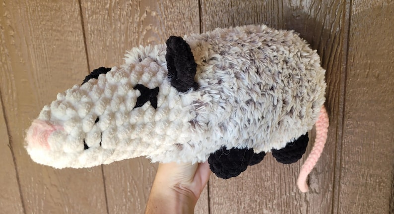Puede incluir: Un juguete de peluche de zarig&uuml;eya hecho a mano. La zarig&uuml;eya es gris y blanca con orejas, patas y marcas oculares negras. Tiene una nariz y una cola rosas. El juguete de peluche est&aacute; hecho de un material suave y texturizado.