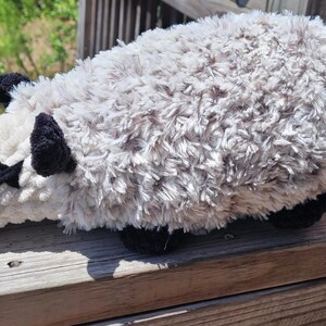 Puede incluir: Un juguete de peluche de zarig&uuml;eya con cabeza blanca, cuerpo gris y cola rosa. La zarig&uuml;eya tiene orejas y pies negros, y una marca negra en forma de estrella en la cara. El juguete est&aacute; hecho de un material suave y texturizado.