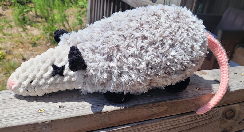 Puede incluir: Un juguete de peluche de zarig&uuml;eya con cabeza blanca, cuerpo gris y cola rosa. La zarig&uuml;eya tiene orejas y pies negros, y una marca negra en forma de estrella en la cara. El juguete est&aacute; hecho de un material suave y texturizado.