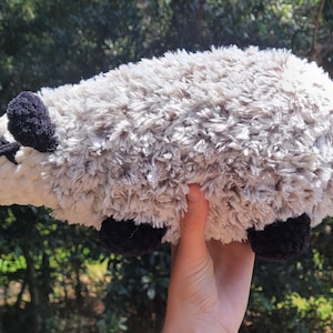 Puede incluir: Un juguete de peluche de zarig&uuml;eya con cabeza blanca, cuerpo gris, orejas y pies negros y cola rosa. El animal de peluche hecho a mano se sostiene contra un fondo verde borroso.