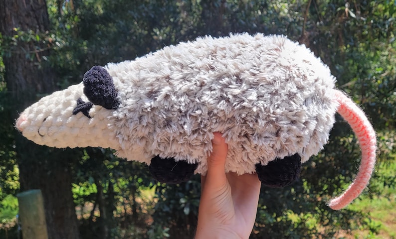 Puede incluir: Un juguete de peluche de zarig&uuml;eya con cabeza blanca, cuerpo gris, orejas y pies negros y cola rosa. El animal de peluche hecho a mano se sostiene contra un fondo verde borroso.