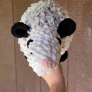 Puede incluir: Un juguete de peluche de zarig&uuml;eya hecho a mano con un cuerpo blanco, pelaje gris en la parte superior y orejas negras. La zarig&uuml;eya tiene una nariz rosa y ojos negros peque&ntilde;os. El juguete est&aacute; hecho de hilo suave y texturizado.