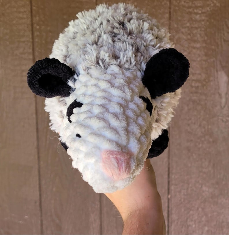 Puede incluir: Un juguete de peluche de zarig&uuml;eya hecho a mano con un cuerpo blanco, pelaje gris en la parte superior y orejas negras. La zarig&uuml;eya tiene una nariz rosa y ojos negros peque&ntilde;os. El juguete est&aacute; hecho de hilo suave y texturizado.