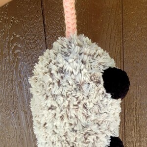 Puede incluir: Un juguete de zarig&uuml;eya de peluche hecho a mano con un cuerpo gris y blanco esponjoso, orejas negras y una cola rosa. El juguete se sostiene por un asa rosa de ganchillo.