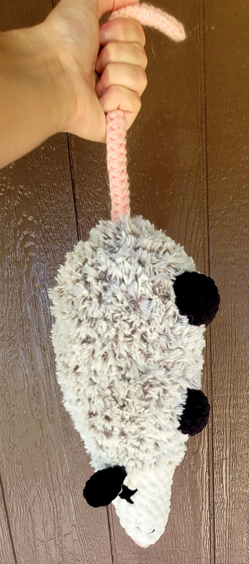 Puede incluir: Un juguete de zarig&uuml;eya de peluche hecho a mano con un cuerpo gris y blanco esponjoso, orejas negras y una cola rosa. El juguete se sostiene por un asa rosa de ganchillo.
