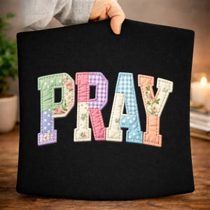 以下が含まれることがあります： 黒いクッションに「PRAY」の文字がカラフルなパッチワークでアップリケされています。文字は、ピンク、紫、青、緑の色合いで、花柄、水玉模様、ギンガムチェック柄です。