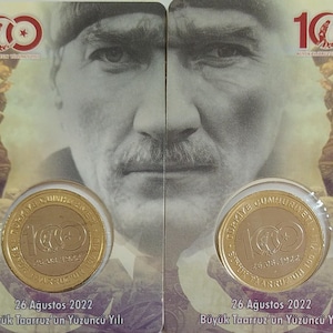 Op de afbeelding: Twee gouden herdenkingsmunten met de tekst "TURKIYE CUMHURIYETI" en "100. YIL" op de voorkant. De munten zijn omgeven door een rood-wit-sterrenontwerp. De tekst "26 Agustos 2022" en "Buyuk Taarruz'un Yuzuncu Yili" staat onder de munten.