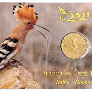 May include: A gold coin featuring a hoopoe bird with a long beak and a crest of feathers. The coin is in a plastic case with a picture of a hoopoe bird and the text "Anadolu'ya Özgü Kuşlar İbibik / Hoopoe" and "2021".