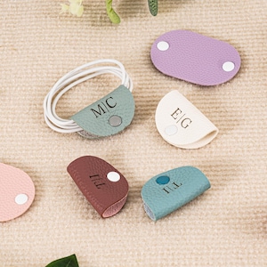 Puede incluir: Una colección de organizadores de cables personalizados en varios colores, incluyendo rosa, morado, verde azulado y marrón. Cada organizador presenta iniciales y un cierre de botón a presión. Un cable blanco está adjunto.