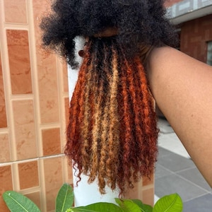 May include: A bundle of hair extensions featuring a dark black base at the top that transitions into a vibrant mix of copper, auburn, and golden blonde tones. The hair is styled in textured, coiled locs with loose, curly ends throughout the length.