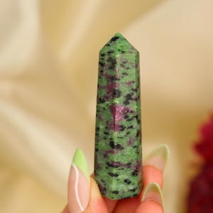 May include: A green and red zoisite crystal tower. The polished stone has a rectangular shape with a pointed top. The crystal is a mix of green, black, and red colours. The background is a soft, blurred beige.
