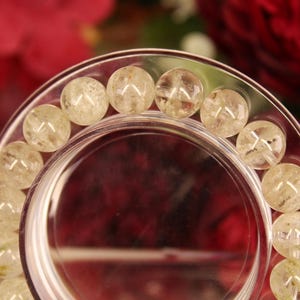 May include: Close-up of a clear glass bracelet holder with a circular design. The holder displays a bracelet made of round, light-colored beads. The background features blurred red and green floral elements.