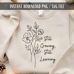 May include: A cream-colored sweatshirt with a black floral design and the text "Still Growing, Still Learning." The design features stylized flowers and leaves. The words are in a simple, elegant font.