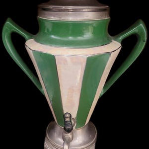 May include: A tall ceramic beverage dispenser featuring alternating vertical stripes of forest green and iridescent cream. The vessel includes two large green side handles, a metallic lid with a finial, and a metallic base with three legs and a spigot.