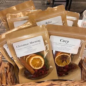 Puede incluir: Varios sobres transparentes llenos de frutas y especias secas, como naranjas, arándanos y canela. Los sobres están etiquetados con nombres como "Christmas Morning" y "Cozy" y se presentan en una cesta tejida.