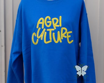 May include: A royal blue sweatshirt with the word "AGRICULTURE" in yellow, stylized lettering. A white and light blue butterfly graphic is on the lower right side. The sweatshirt is displayed on a white mannequin.