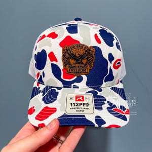 May include: A five-panel baseball cap featuring a red, white, and blue camouflage pattern. A brown leather patch on the front displays an eagle emblem with the text 250 YEARS OF FREEDOM 1776-2026. A white sticker on the brim reads EST. 1970 112PFP PRINTED 5 PANEL OSFM.