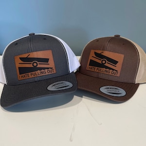 Könnte beinhalten: Zwei Trucker-Caps mit einem braunen Leder-Patch, auf dem "Ich hasse es, rauszuziehen" steht, und einer Silhouette eines Bootes, das aus dem Wasser gezogen wird. Die Mützen sind grau und braun mit einem weißen Netzrücken.