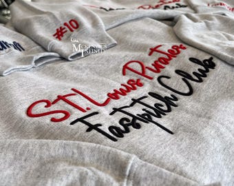 May include: Multiple light grey sweatshirts with embroidered text. One sleeve shows the number "#10" in red. The sweatshirts feature the text "STL Louis Pirates Fastpitch Club" in red and black embroidery. The McKenzie's Infinity logo is also visible.