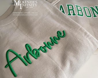 May include: Two beige sweatshirts with green embroidered lettering. One sweatshirt says "Arbonne" in cursive, and the other says "ARBONNE" in block letters with a white outline. The sweatshirts are on a white surface.