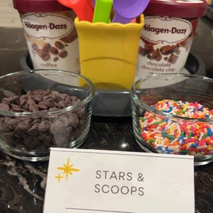 May include: An ice cream station featuring two Häagen-Dazs pints in coffee and chocolate chocolate chip flavors, a yellow holder with red, green, and purple spoons, two glass bowls with chocolate chips and sprinkles, and a sign reading STARS & SCOOPS.
