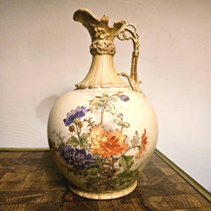 May include: A cream-colored ceramic pitcher featuring a hand-painted floral design with orange, yellow, and purple chrysanthemums. The vessel has a fluted neck, a decorative gold-toned handle, and intricate gold detailing around the rim and base.