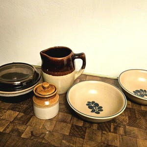 Op de afbeelding: Een verzameling vintage steengoed keukengerei, waaronder een ovenschotel met deksel, een kleine pot met deksel, een kan en twee kommen. De kommen hebben een beige interieur met een blauw bloemmotief en een groene rand. De items staan op een houten oppervlak.