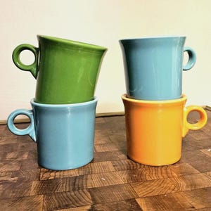May include: Four ceramic mugs in different colours. The mugs are stacked in pairs. The top mug is green, the second mug is blue, the third mug is orange, and the bottom mug is blue.