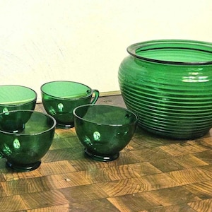 May include: A collection of green glass drinkware, including four teacups with handles and a large, round bowl. The bowl has horizontal ridges. The items are arranged on a wooden surface.