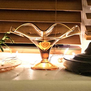 May include: A translucent amber-colored glass compote dish featuring a wide, ruffled, scalloped rim and a pedestal base. The bowl has a smooth, curved silhouette that tapers down to a circular foot, reflecting warm light through its glass surface.