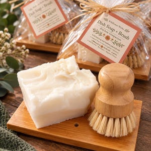 May include: A white rectangular block of dish soap and a round bamboo palm brush with light-colored bristles resting on a rectangular wooden tray. The soap has a textured surface and the brush features a smooth, light-brown wooden handle.