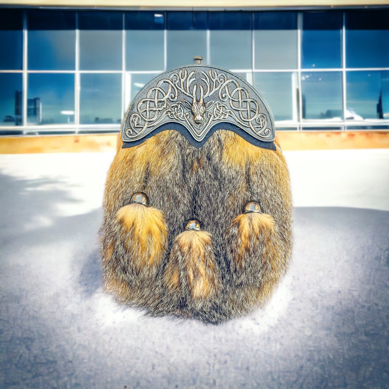 May include: A traditional Scottish sporran, a fur pouch, rests on a snowy surface. The sporran features a decorative silver metal cantle with Celtic knotwork and a central animal figure. The fur is a mix of brown, gray, and golden tones, with three decorative tassels.