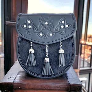 May include: Black leather sporran with a Celtic knot design and silver-toned accents. The flap features embossed knotwork and is embellished with small, round, silver-colored studs. Three black tassels hang from the bottom, adding a decorative touch.
