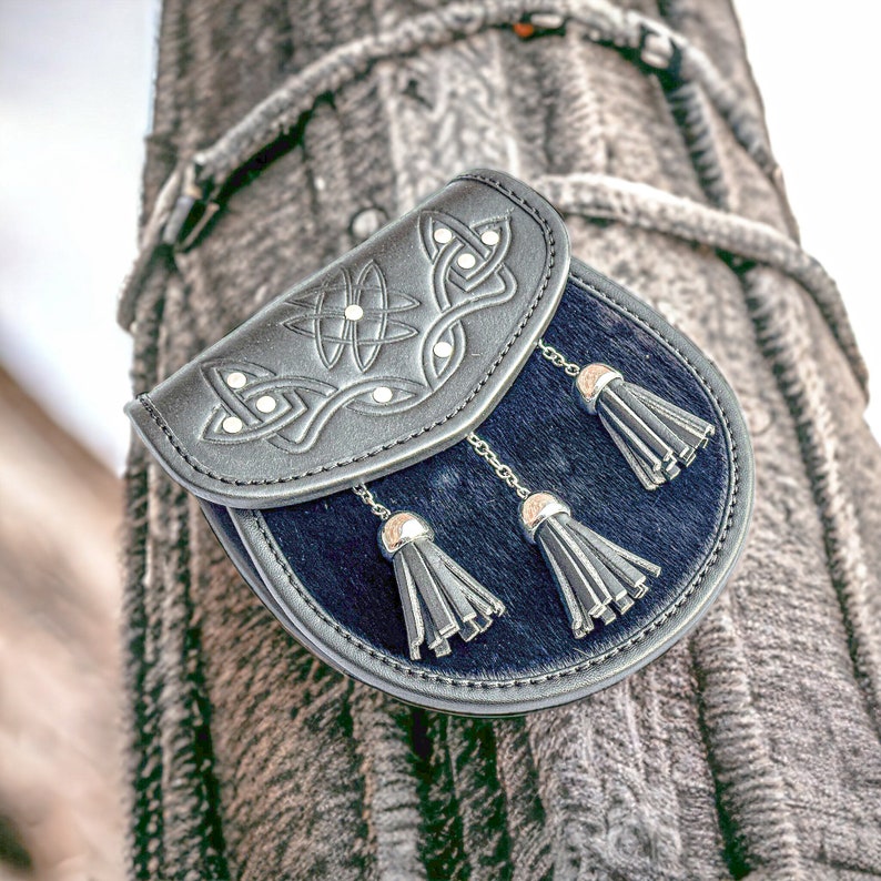 May include: A black leather sporran with Celtic knotwork design on the flap. The sporran has a dark blue fur body, silver chain tassels, and silver caps. This traditional Scottish accessory is designed to be worn with a kilt.