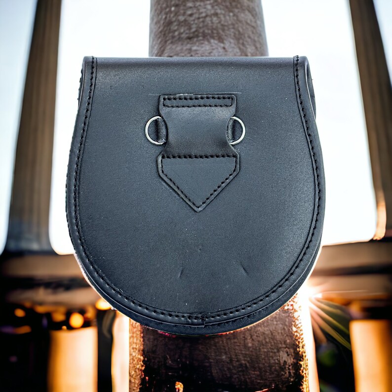 May include: A black leather sporran, a traditional Scottish pouch, with a rounded bottom and a flap closure. The sporran has a decorative stitched border and silver-colored metal loops for attaching a chain or strap. The background is blurred.